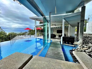 A modern infinity pool with a clear view of the ocean, a covered seating area with red umbrellas, and a stone-tiled deck. - Casa Ventanas: Stunning new Architectural Gem! (Manuel Antonio)