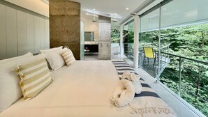 Un dormitorio moderno con una cama grande, vistas a zonas verdes a través de amplios ventanales, y un baño visible al fondo.