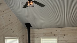 A cozy living room with a fireplace, two sofas, a red chair, and a ceiling fan. - Super clean cabin near ORV trails & AuSable River, walking distance to town Mio. (Mio)