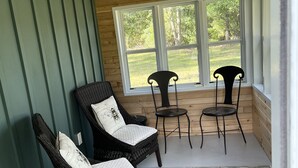A cozy room with wooden walls and ceiling, featuring two wicker chairs with cushions, a small table, and a large window offering a view of greenery outside. - Super clean cabin near ORV trails & AuSable River, walking distance to town Mio. (Mio)