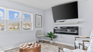 A living room with a flat-screen TV, a large window with blinds, a cushioned ottoman, a patterned rug, and a decorative chair. - Lovely 2-Bedroom Condo, Close to Burlington/Airport! (South Burlington)