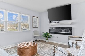 A living room with a flat-screen TV, a large window with blinds, a cushioned ottoman, a patterned rug, and a decorative chair.