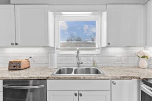 A kitchen with white cabinets, a stainless steel sink, a window with blinds, a toaster, and a vase with flowers. - Lovely 2-Bedroom Condo, Close to Burlington/Airport! (South Burlington)