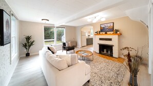 Ein geräumiges Wohnzimmer mit einem weißen Sofa, einem Couchtisch und einem Kamin. Es gibt einen Essbereich mit Tisch und Stühlen, und im Hintergrund eine Küche.