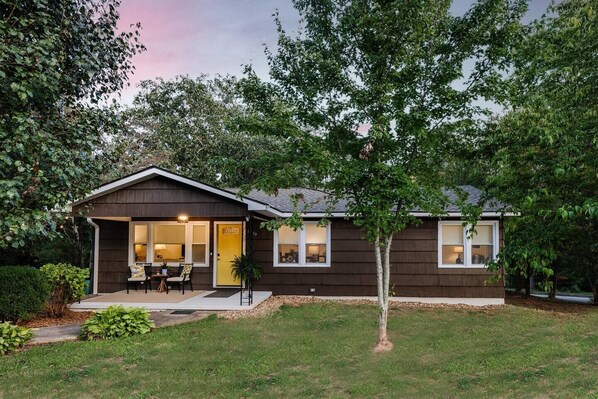 A single-story house with a covered porch, a small garden, and a tree in front.