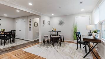 A dining area with a round table, chairs, and a rug. There are doors and a window with a plant on the right.