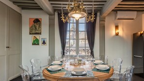 Un comedor con una mesa redonda preparada para seis comensales, sillas transparentes, una araña, obras de arte en las paredes y vistas a un edificio a través de una ventana.