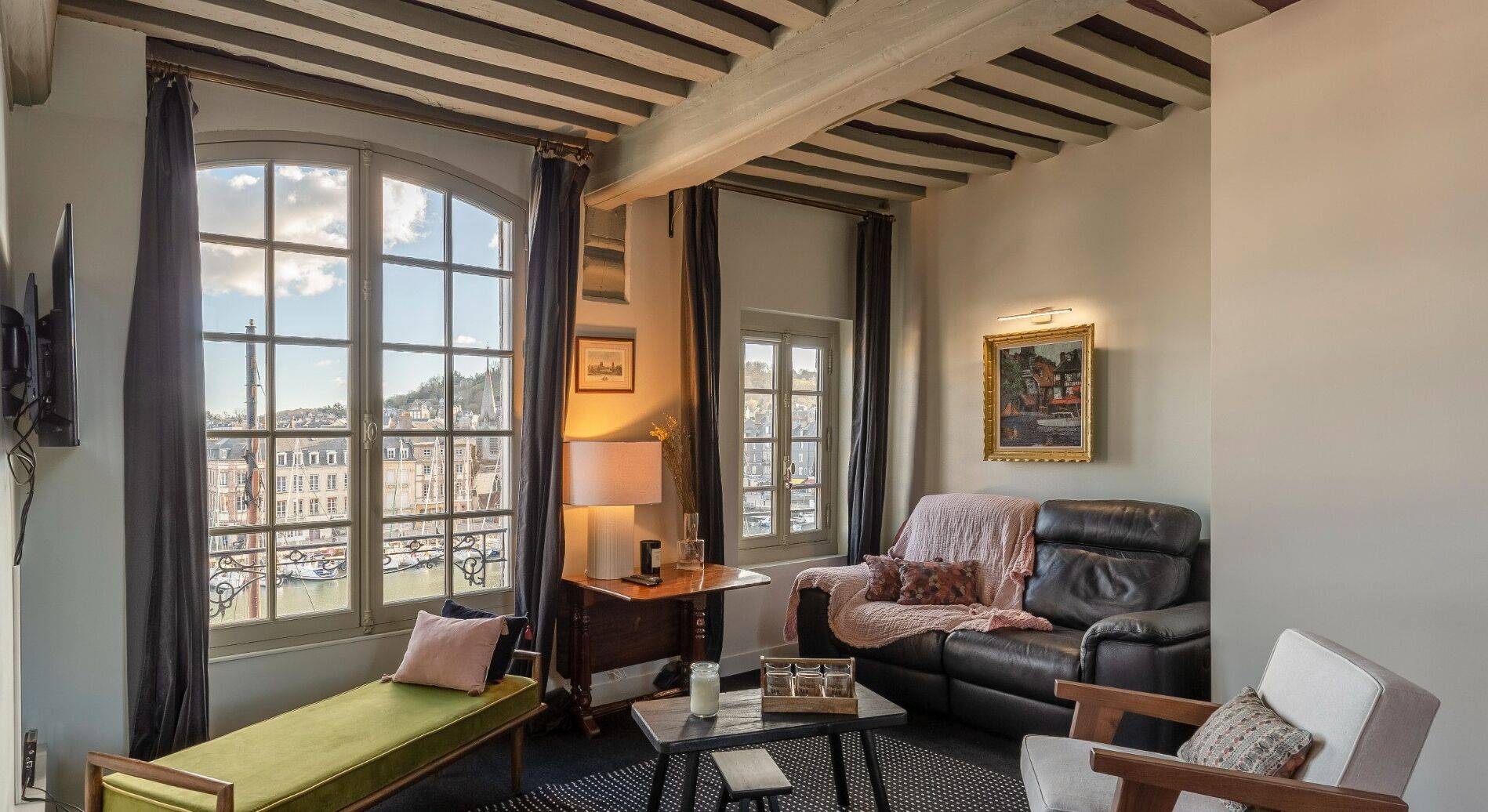 A cozy room with a view of a harbor, featuring a green bench, a leather armchair, a small table, and a lamp.