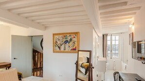 Una habitación con una cama, un cuadro, un espejo y una ventana con vistas a edificios.