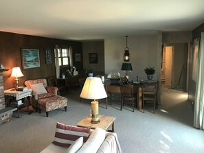 Una sala de estar acogedora con sofá, sillón y lámpara de mesa. Se observa un comedor con mesa y sillas al fondo.