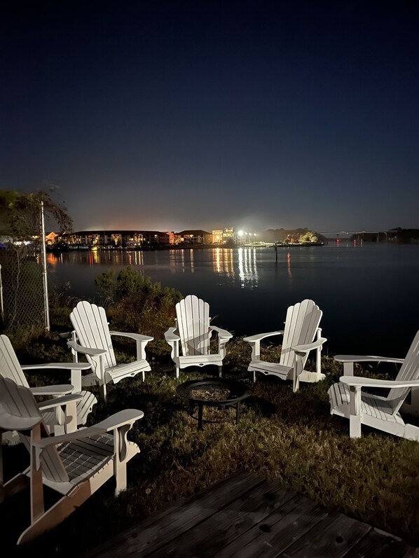 A lakeside deck with white Adirondack chairs and a fire pit, overlooking a calm lake with a distant illuminated bridge.