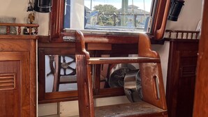 A wooden cabin interior with a steering wheel, a fan, and a view of the outside through a window.