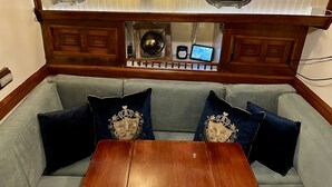 A cabin interior with a wooden ceiling, two porthole windows, a wooden bench, and a cushioned seating area with decorative pillows.