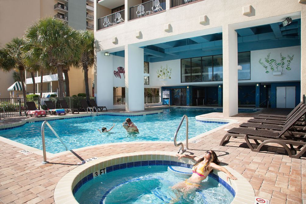 A swimming pool area with lounge chairs, palm trees, and a building with balconies.
