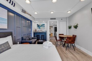 A hotel room with a bed, a sofa, a dining table with chairs, a kitchen area, and a hallway leading to another room.