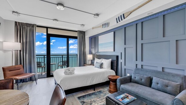 A hotel room with a bed, a sofa, a dining table, and a view of the ocean through the balcony doors. - Breakers Tower 1BR Oceanfront Condo (Myrtle Beach)