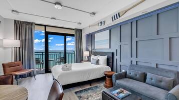 A hotel room with a bed, a sofa, a dining table, and a view of the ocean through the balcony doors.