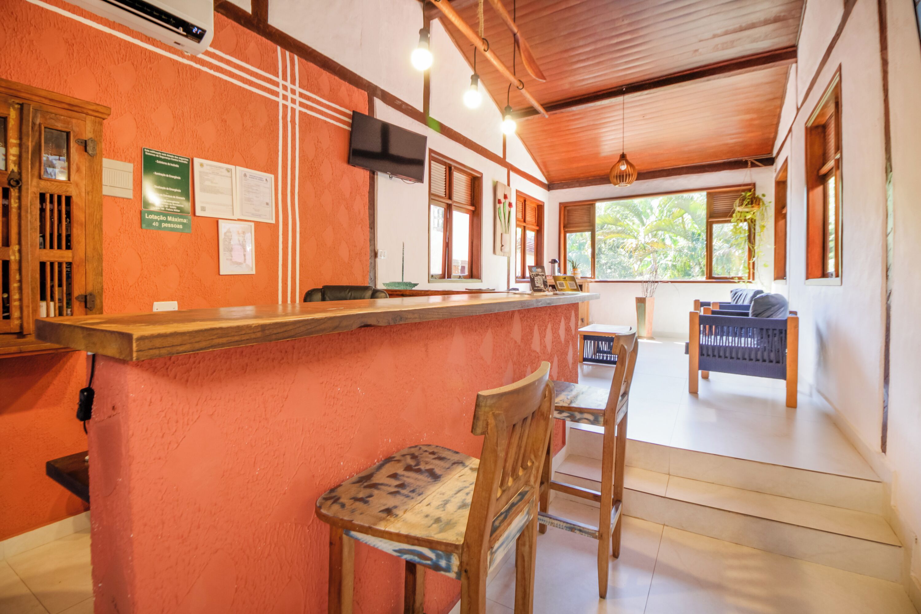 A room with a wooden counter and chairs, a television mounted on the wall, and a window with a view of greenery outside.