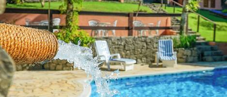 A pool with clear blue water, surrounded by stone tiles and a stone wall. There are lounge chairs and a small grassy area in the background.