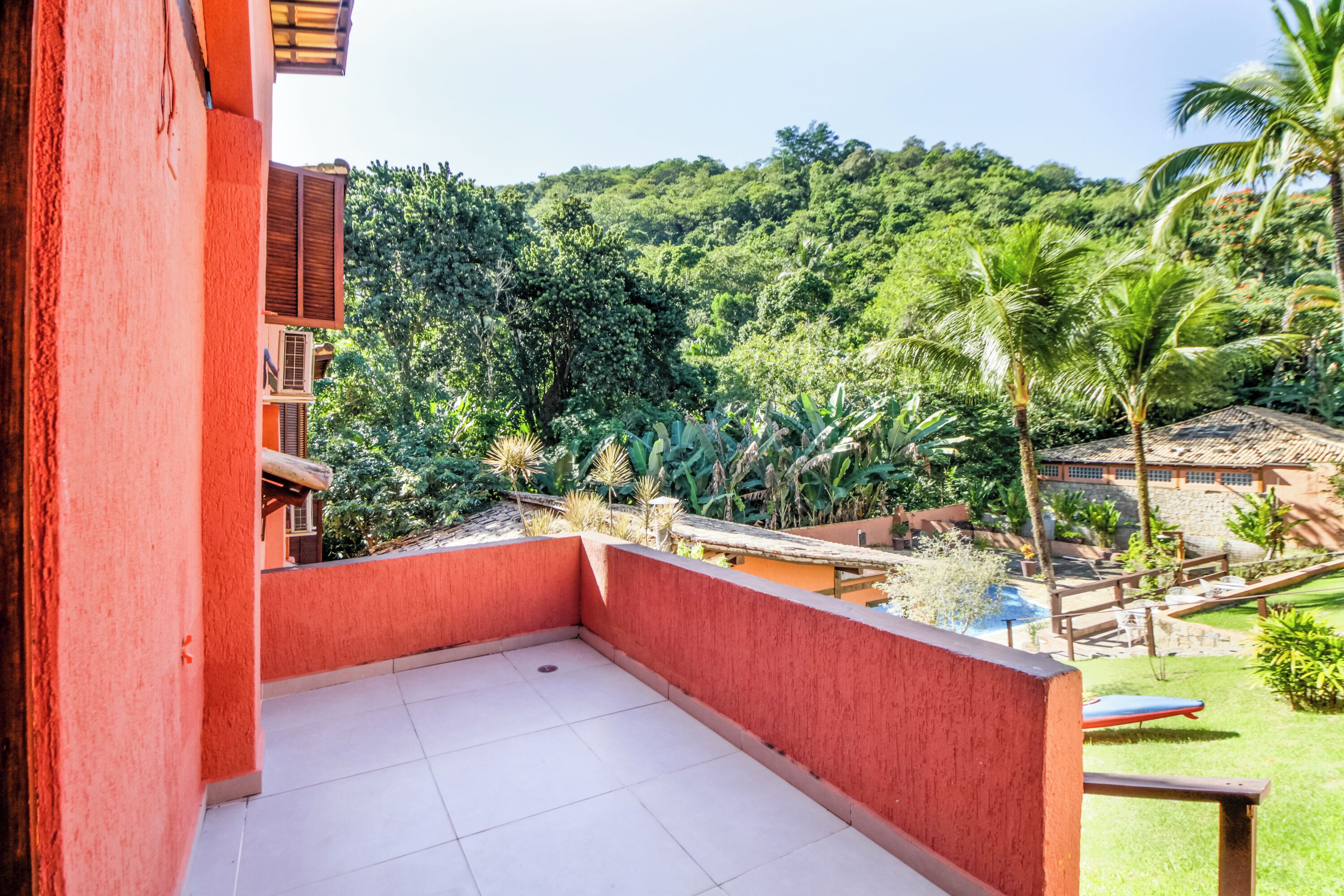 A red building with a tiled balcony overlooking a lush garden with palm trees and a pool area.