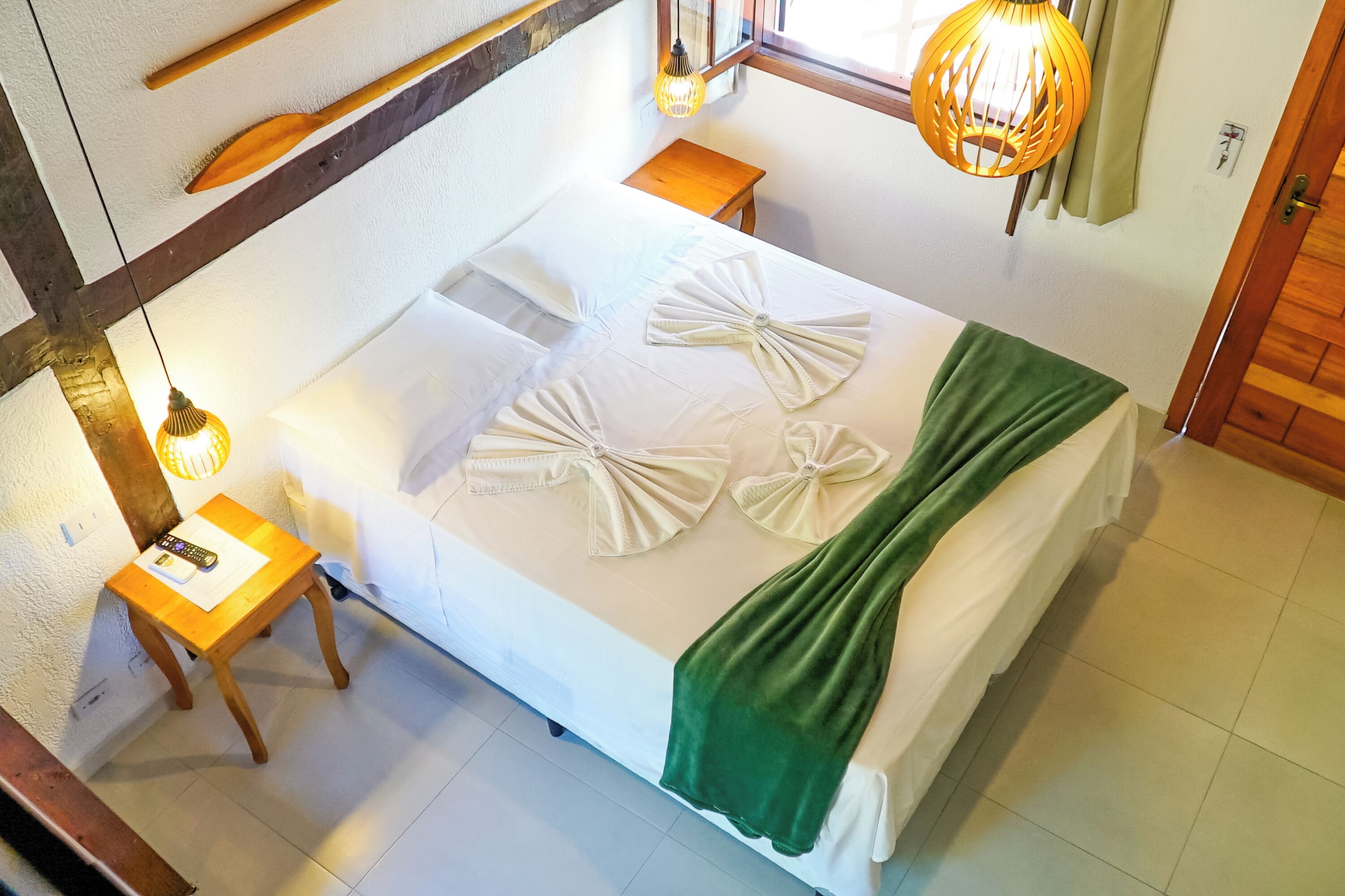 A neatly made bed with towels arranged in bows, a wooden bedside table with a remote control, a small wooden stool, and a hanging lamp.
