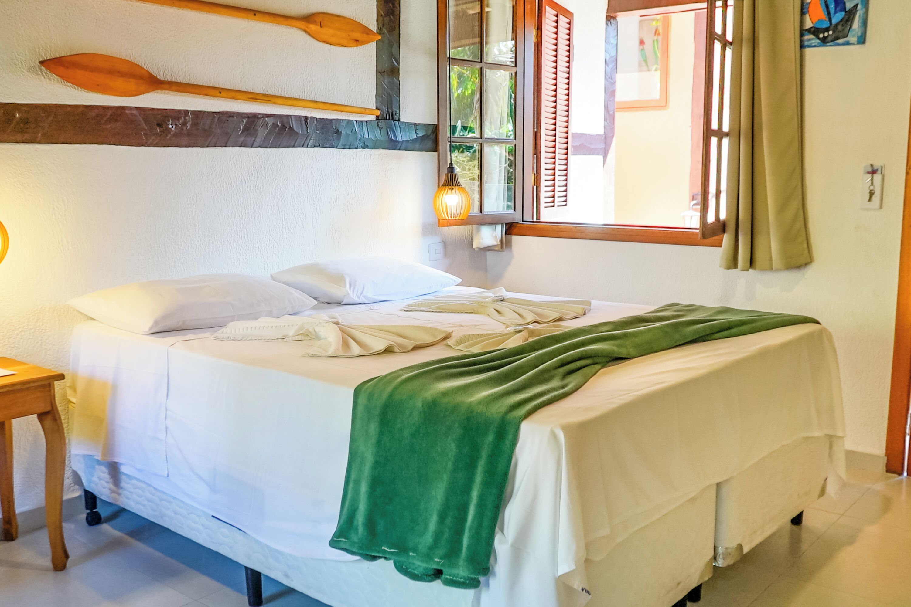 Una cama bien tendida con una manta verde, adornos de remo de madera en la pared, una lámpara de noche y una ventana con contraventanas.