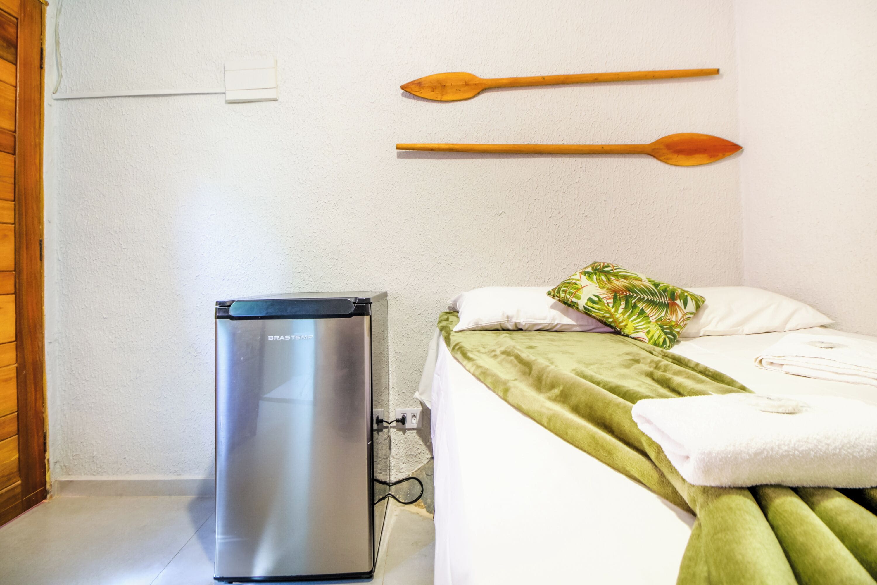 Una habitación compacta con cama, un refrigerador pequeño y dos cucharas de madera colgadas en la pared.