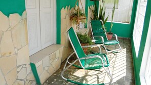 A covered porch with two green chairs, a stone wall, and a white door. - Hostal Rodolfo y Nancy TRINIDAD (Trinidad)