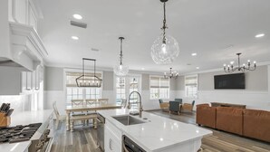 Una cocina amplia con una isla central, un comedor con mesa y sillas, una sala de estar con sofá, y un televisor grande montado en la pared.
