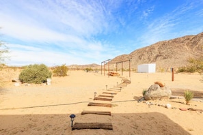 A desert landscape with a wooden pathway leading to a structure with a metal frame and a white sign. - The GOAT Desert Experience (Landers)