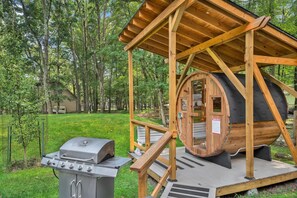 Een houten sauna met een deur in de vorm van een vat en een barbecue ervoor.