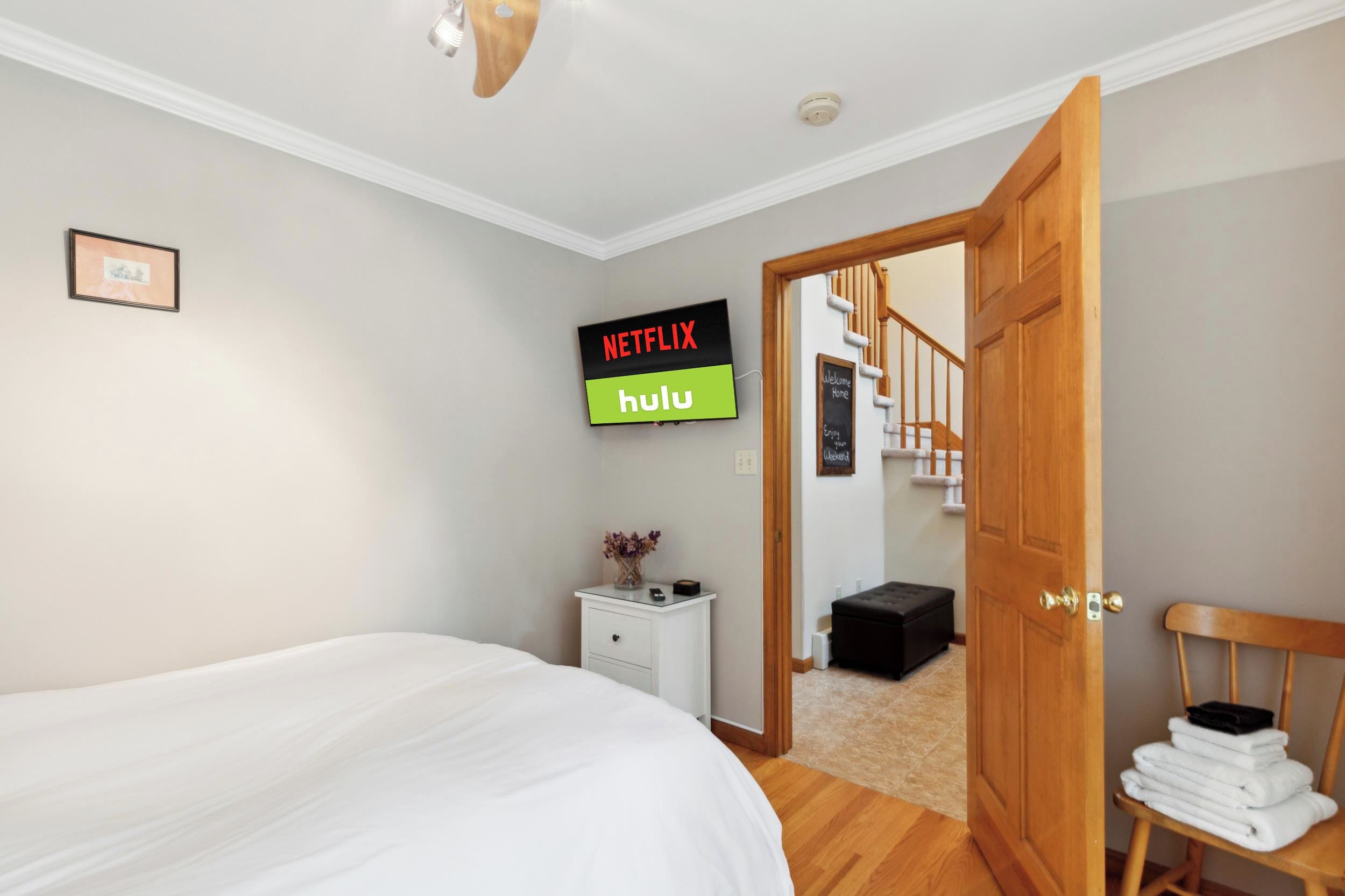 A bedroom with a bed, a nightstand with a vase, a sign for Netflix and Hulu, and a wooden door leading to another room.
