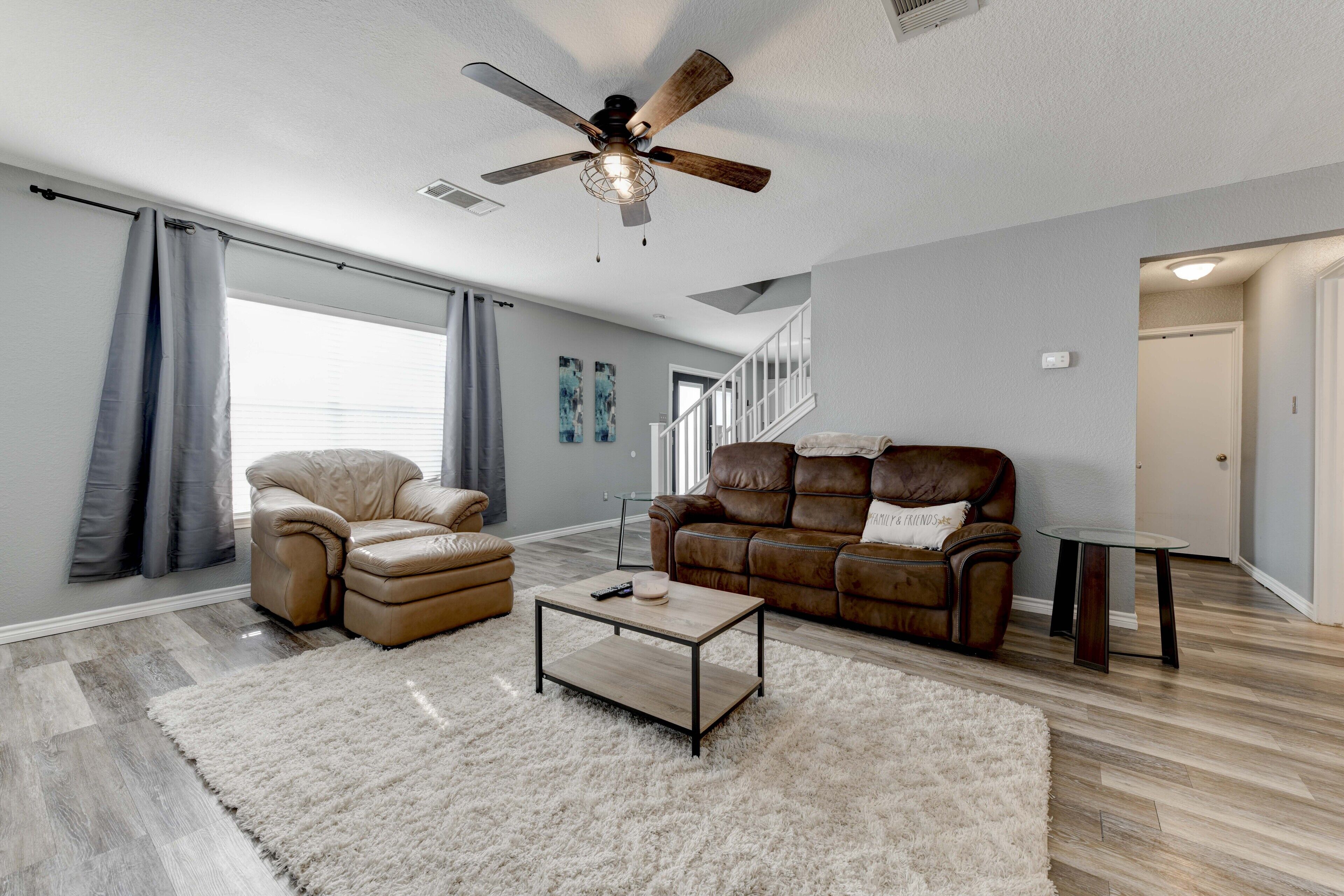 A spacious living room with a ceiling fan, leather sofas, a coffee table, and a large window with curtains.