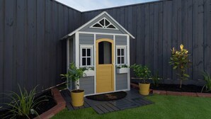 Una casita de juegos de madera con puerta amarilla, detalles blancos y techo triangular. Está rodeada por una cerca oscura y tiene dos macetas con plantas a cada lado.