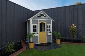 A small wooden playhouse with a yellow door, white trim, and a triangular roof. It is surrounded by a dark fence and has two potted plants on either side.