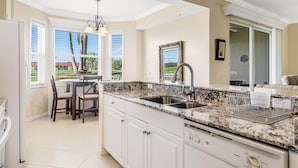 A kitchen with white cabinets, granite countertops, a double sink, and a dining area with chairs. - Golf Membership Available! 2 BR/2BA, Pickleball, Pools, Near Gulf Beaches (Naples)