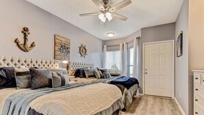 Un dormitorio con cabecera capitonada, dos camas con ropa de cama a juego, ventilador de techo y decoración de paredes con temática náutica.