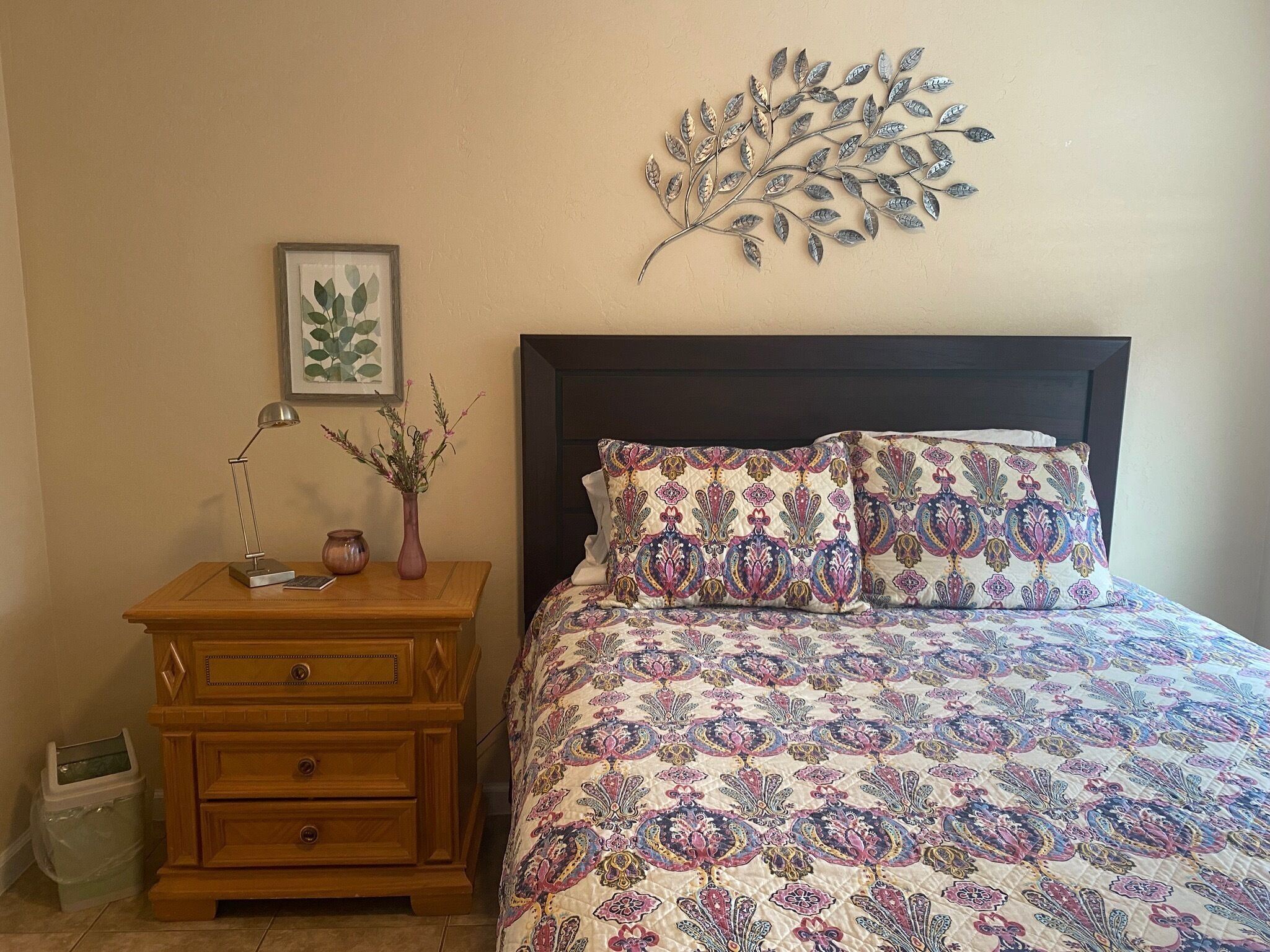 Un dormitorio con una cama, una mesita de noche, una lámpara, un jarrón con flores y una obra de arte en la pared.