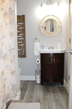 A bathroom with a wooden floor, a round mirror, a white sink, and a wooden fish decoration on the wall. - Nature's Calling at Port Oliver (Scottsville)