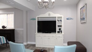 Una sala de estar con un televisor sobre un mueble blanco, dos sillones azul claro, un reposapiés marrón y una araña.