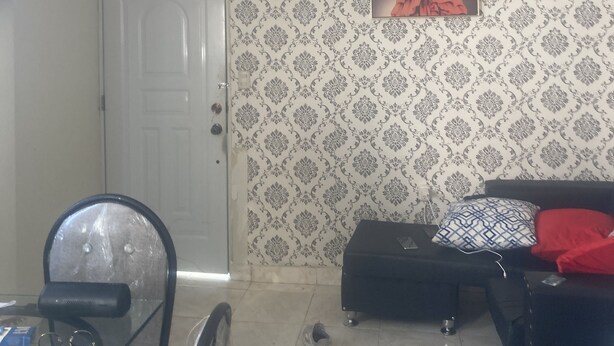 Habitación con ventilador de techo, papel tapiz estampado, una puerta con un letrero, un cuadro enmarcado, un sofá negro con un cojín rojo y una mesa de vidrio con una silla.