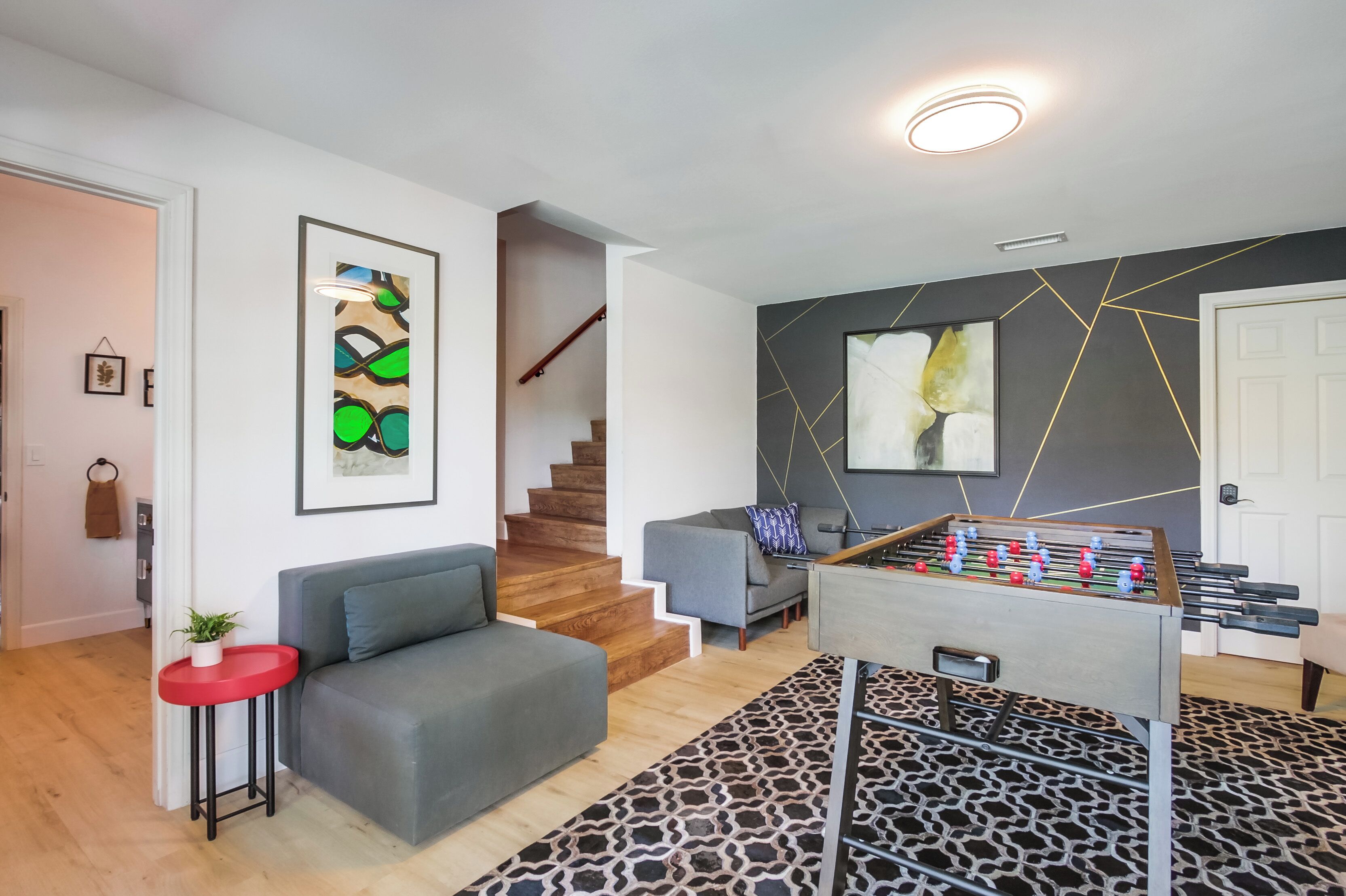 A modern living room with a grey sofa, a red side table with a plant, a foosball table, and abstract wall art.