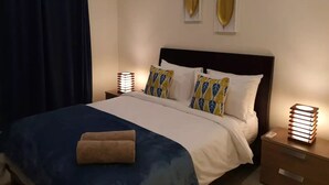 A neatly made bed with a blue blanket and a rolled towel, two bedside lamps, and an air conditioning unit on the wall. - Marigold Apartment 4.  2 bedroom 2nd floor apartment with a great view. (Nadi)