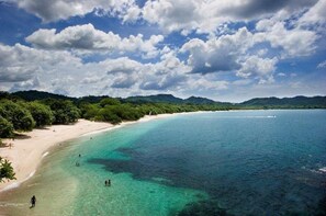 A beach with clear turquoise waters, a sandy shore, and a forested hill in the background.
