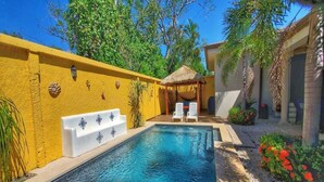 Ein Pool mit gelber Wand, einem Gazebo mit Reetdach und weißen Liegestühlen.
