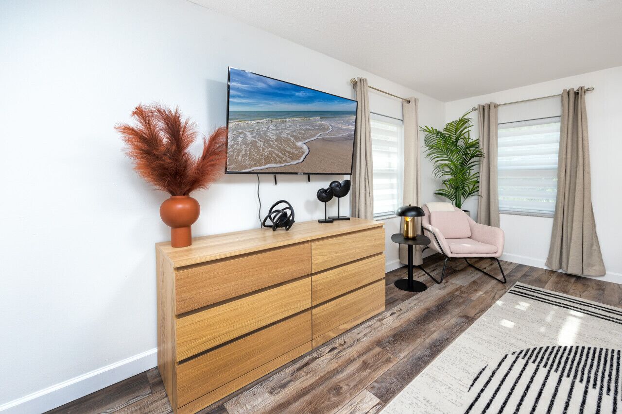 Una sala de estar moderna con un mueble de madera, un televisor montado en la pared, un sillón rosa y un ventanal con cortinas.