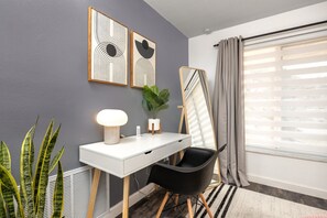 Un dormitorio moderno con un escritorio blanco, una silla negra, un espejo grande y una planta en maceta.