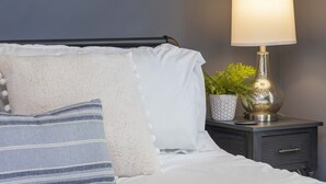A neatly made bed with white and blue striped pillows, a fluffy white pillow, and a small potted plant on a bedside table.