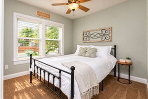A bedroom with a bed, a ceiling fan, a window with blinds, a wall-mounted artwork, and a small bedside table with a plant.