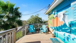 Una terraza de madera con dos sillas Adirondack azules y una pequeña mesa, un mural de un ave en el edificio, y una palmera al fondo.